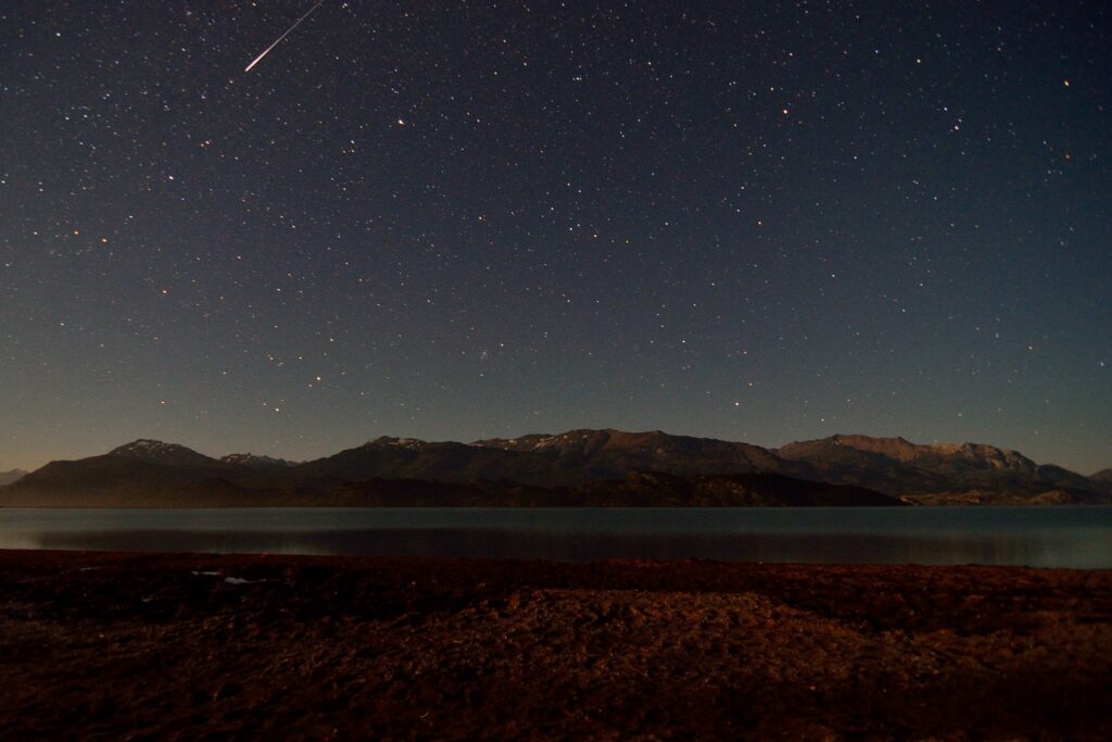 Starry night sky over mountains and serene lake with a shooting star.